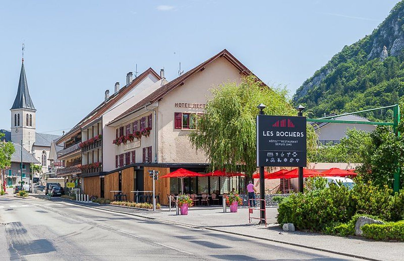 Façade de l'hôtel-restaurant Les Rochers, terrasse aux parasols rouges et clocher du village