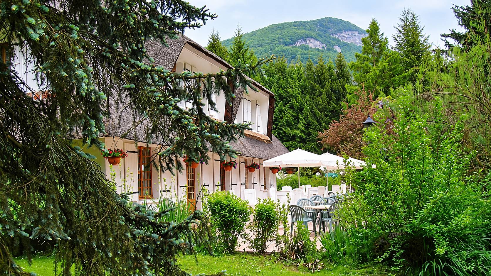 La Chrissandière, maison au toit de chaume et sa terrasse fleurie au pied d’une montagne